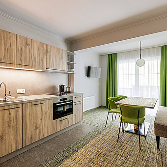 Neben einer modernen Küche steht ein Esstisch mit grünen Stühlen.