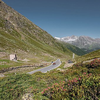Ein rotes Auto fährt auf einer Landstraße durch eine Bergkulisse.